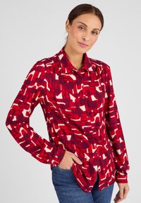 Camicia a maniche lunghe con pattern rosso e crema, dotata di collo e vestibilità rilassata. Tessuto morbido.