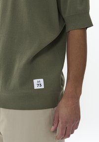 Tričtvrteční, olivově zelený svetr s texturovaným pletením, s bílou nálepkou s nápisem "M 73" na levé straně blízko lemu.