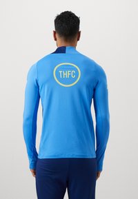 Blå langærmet skjorte med et rundt gult logo med "THFC" på ryggen, lavet af letvægtsstof med en glat tekstur.