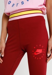 Červené sportovní legíny s elastickým pasem, které mají růžové a žluté pruhy. Obsahují červené logo Nike a text "SPORTOVNÍ OBLEČENÍ".