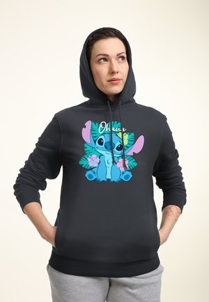 Frau trägt einen dunklen Hoodie mit einem bunten Cartoon-Charakter und tropischen Blättern auf der Vorderseite, Hände in den Taschen, schaut leicht nach oben.