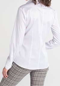 Chemise blanche à boutons avec manches longues, coupe ajustée, détail de couture au dos. Associée à un pantalon à motif écossais dans des tons neutres.
