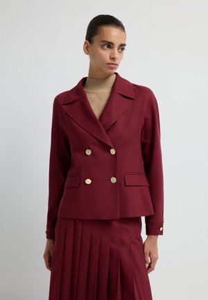 Donna che indossa un blazer bordeaux doppiopetto con bottoni dorati e una gonna plissettata abbinata su un maglione a dolcevita beige.
