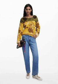 Džemper pleteni uzorak s cvjetnim motivima u žutoj i smeđoj boji, upareno sa svijetloplavim trapericama ravnih nogavica. Model drži malu crnu torbu.