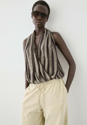 Jong persoon met donkere zonnebril, mouwloze bruin-beige gestreepte top en lichtbeige losse broek met koordzakken.