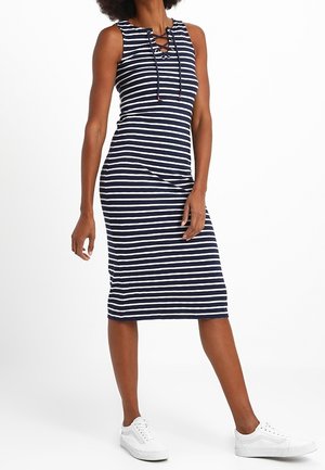 Femme portant une robe sans manches rayée marine et blanche, longueur genou, avec un col lacé et des baskets blanches, debout sur fond blanc.