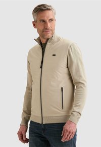 Homme aux cheveux gris courts portant un gilet beige zippé par-dessus une chemise à manches longues, debout et regardant sur le côté devant un fond uni.