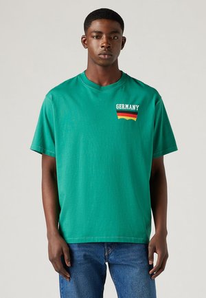 Grøn bomulds T-shirt med ordet "TYSKLAND" i hvidt, med et design i røde, sorte og gule striber nedenunder. Rund hals og korte ærmer.
