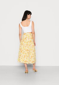 Top crop sem mangas branco combinado com uma saia midi amarela com flores, apresentando margaridas. A saia tem uma cintura franzida e flui de forma solta. Sandálias de salto amarelas.