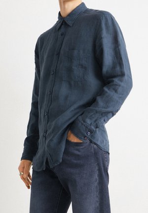 Mand iført mørkeblå skjorte med knapper, lange ærmer og frontlomme samt mørkeblå jeans, hånd i lommen mod ensfarvet hvid baggrund.