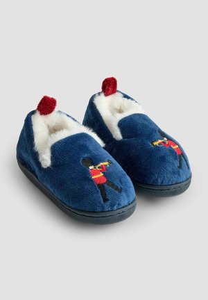Sinine pehme susside paar, valge fliisvoodriga ja punaste pom-pomidega, küljes on tikand, kus on kujutatud valvurit, kes mängib trompetit.