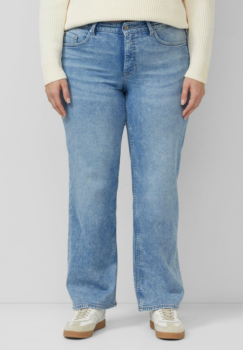 Person, die hellblaue Jeans mit geradem Bein, weiße Sneakers und einen cremefarbenen, gerippten Pullover trägt und vor einem schlichten Hintergrund steht.