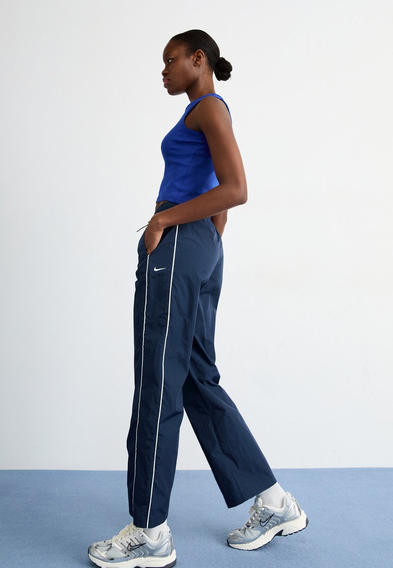 Marine blauwe trainingsbroek met witte zijstripes, gecombineerd met een blauwe mouwloze top en zilveren sportschoenen. Model poseert zijwaarts.