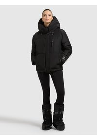 Mujer con chaqueta acolchada negra de cuello alto, leggings negros y botas de invierno negras, de pie contra un fondo liso y claro.