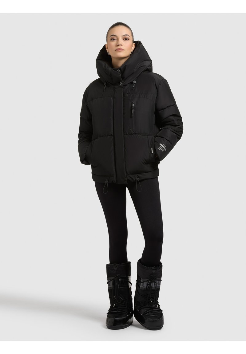 Mujer con chaqueta acolchada negra de cuello alto, leggings negros y botas de invierno negras, de pie contra un fondo liso y claro.