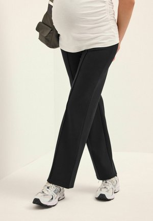 Schwangere Person, die ein weißes gerafftes Oberteil, schwarze Hosen, weiße Turnschuhe und eine schwarze Umhängetasche trägt und mit überkreuzten Beinen steht.