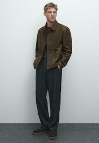 Braune Lederjacke mit strukturiertem Design, dunkelgraue Hose und dunkle Schuhe, vor einem hellen Hintergrund mit matter Oberfläche.