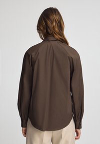 Camicia marrone a maniche lunghe con colletto, caratterizzata da un orlo arrotondato e polsini con bottoni. Texture liscia, vestibilità aderente, vista posteriore.