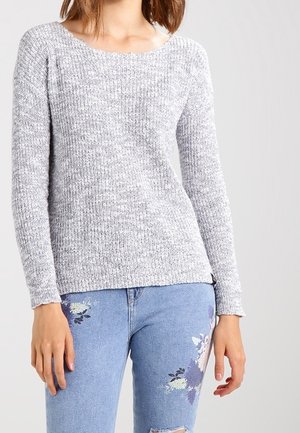 Hellgrauer Strickpullover mit weitem Halsausschnitt, langen Ärmeln und gerippten Bündchen, kombiniert mit ausgewaschenen blauen Jeans mit floralen Mustern.