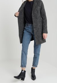 Cappotto lungo in misto lana grigio con rever a punta, due tasche frontali, abbinato a jeans a vita alta blu e stivaletti neri con finitura lucida.
