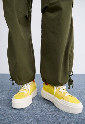 Kollase värvi sneakersid, millel on valged sissepääsud ja paelad, valmistatud tekstuurilisest materjalist. Koos vabas vormis oliivroheliste, elastneäärtega püksidega.
