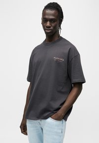 Σκούρο γκρι βαμβακερό t-shirt με κοντά μανίκια, στρογγυλή λαιμόκοψη και διακριτικό λογότυπο «ALLSAINTS» σε κοραλί. Φορεμένο με ανοιχτόχρωμο μπλε τζιν.