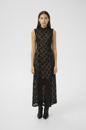 Woman standing wearing a sleeveless black lace maxi dress over a nude underlay with black pointed-toe boots on a plain background.