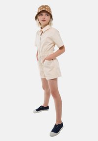 Enfant blond portant une combinaison beige à manches courtes avec fermeture éclair, un chapeau seau à pois marron et des chaussures bateau marines, se tenant les mains dans les poches.