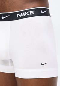 Λευκά ανδρικά εσώρουχα μπόξερ Nike με μαύρη ελαστική ζώνη μέσης που φέρει το λογότυπο Nike και το σήμα swoosh στο πόδι, φορεμένα από ένα άτομο με σκούρο δέρμα.