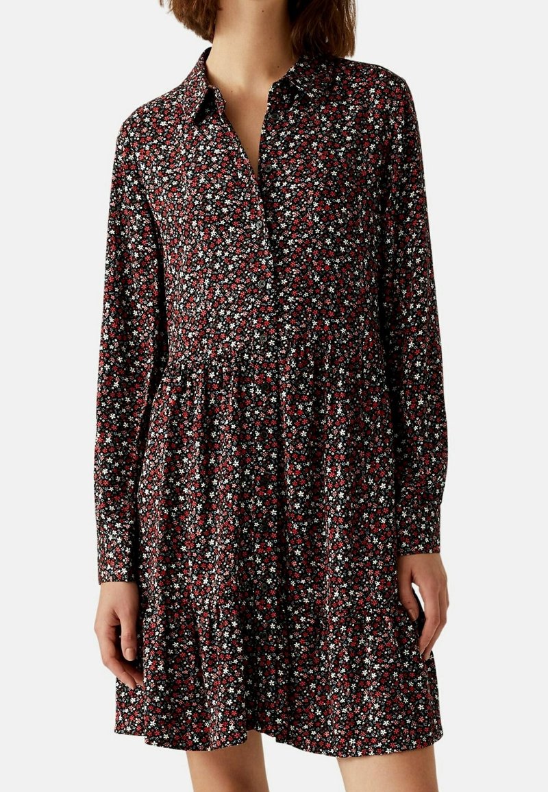 Femme portant une robe à manches longues boutonnée avec un fond noir et un petit motif floral rouge et blanc.