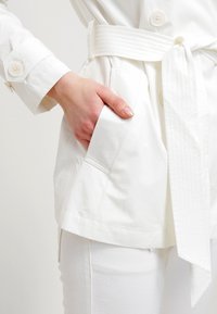 Vita bälte jacka med en slät yta, med sidofickor och dubbelknäppt front, ihopparad med vita byxor.