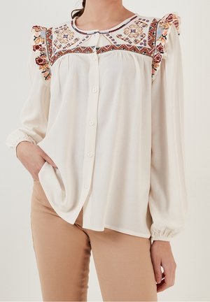 Kobieta ubrana w białą bluzkę z długim rękawem, ozdobioną kolorowym haftowanym karczkiem i falbanami na ramionach, w połączeniu z beżowymi spodniami.