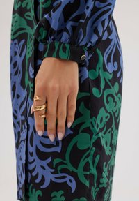 Sukienka z długim rękawem z wyrazistymi niebieskimi i zielonymi wzorami kwiatowymi na czarnym tle. Posiada zapięcie na guziki przy mankiecie oraz złoty pierścień na dłoni.
