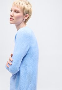 Νεαρό άτομο με κοντά ξανθά μαλλιά, φοράει ασημένια σκουλαρίκια και ανοιχτό γαλάζιο πουλόβερ, κοιτάζει προς τα αριστερά με τα χέρια σταυρωμένα.