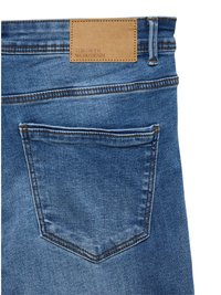 Zilas džinsa bikses ar brūnas ādas etiķeti ar uzrakstu "VERO MODA WE DO DENIM." Aizmugurē redzamas kabatas ar smalku šuvumu un taisnu griezumu.