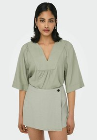 Hellgrüne Bluse mit V-Ausschnitt und gerafften Ärmeln, kombiniert mit einem passenden Wickelrock mit einem schwarzen Knopfdetail.