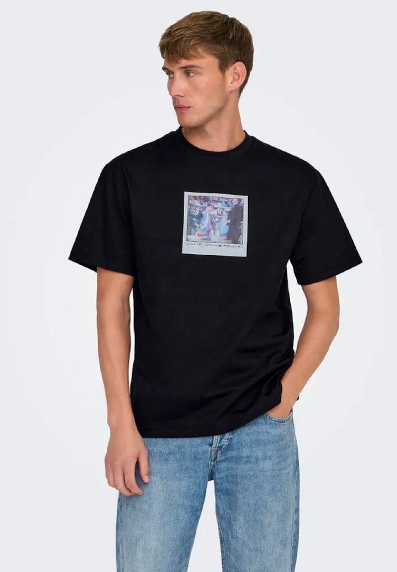 Camisa de algodón negra con mangas cortas, que presenta un estampado gráfico cuadrado en el pecho. Combinada con jeans de color azul claro.