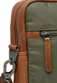 Sac en toile vert avec des accents en cuir marron, comportant une poche zippée et un clip en métal. Coutures visibles le long des bords.