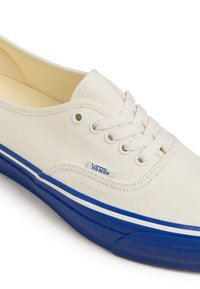 Witte canvas sneakers met een blauwe rubbersole, voorzien van witte veters, een Vans-logo op de zijkant en een klassieke low-top ontwerp.