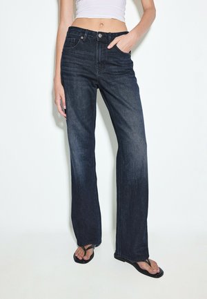 Person iført mørke vidde jeans, hvid cropped top og sorte flip-flops, står med ene hånd i lommen.