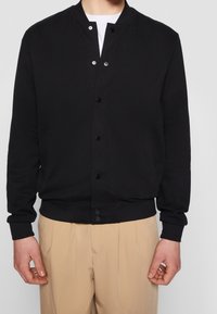 Veste bomber noire en mélange de coton, avec col côtelé, fermeture à boutons et poches latérales. Texture lisse, coupe ajustée.