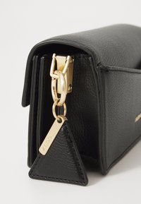 Black textured leather bag with a gold clasp, featuring a triangular tag and sleek design. Accents include a shiny finish and minimalistic style.
