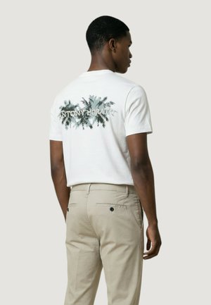 Uomo che indossa una maglietta bianca con una grafica di palme e pantaloni beige chiaro, in piedi con la schiena rivolta verso uno sfondo semplice.