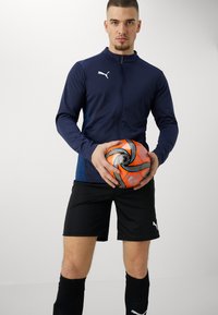 Marineblå atletisk jakke med lynlås, sorte shorts og knælange sokker; holder en struktureret orange og sort fodbold.