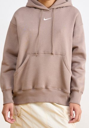 Personne portant un sweat à capuche Nike marron clair avec poche avant et un pantalon couleur crème, visible du cou jusqu'aux hanches.