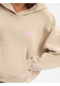 Beige oversized hoodie met een roze 'American Vintage' logo op de borst, voorzien van een voorkantzak en textiel met structuur.