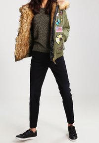 Grön bomberjacka med pälsluva, prydd med olika lappar, tillsammans med en mörk tröja och svarta byxor, avslutad med svarta sneakers.