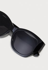 Schwarze Sonnenbrille mit runden Rahmen, glattem Kunststoffmaterial, dunklen Gläsern und auffälligen dicken Bügeln mit einem leichten Akzent an den Schläfen.