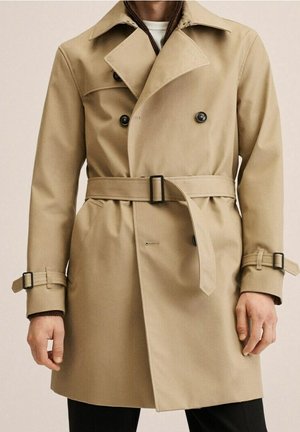 Beiger doppelt geknöpfter Trenchcoat mit schwarzen Knöpfen, Taillengürtel und Manschettenriemen, getragen über dunkler Hose und weißem Unterhemd.