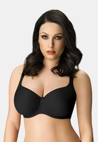 Donna con capelli scuri e mossi indossa un reggiseno nero con ferretti e spalline larghe su uno sfondo chiaro e semplice.